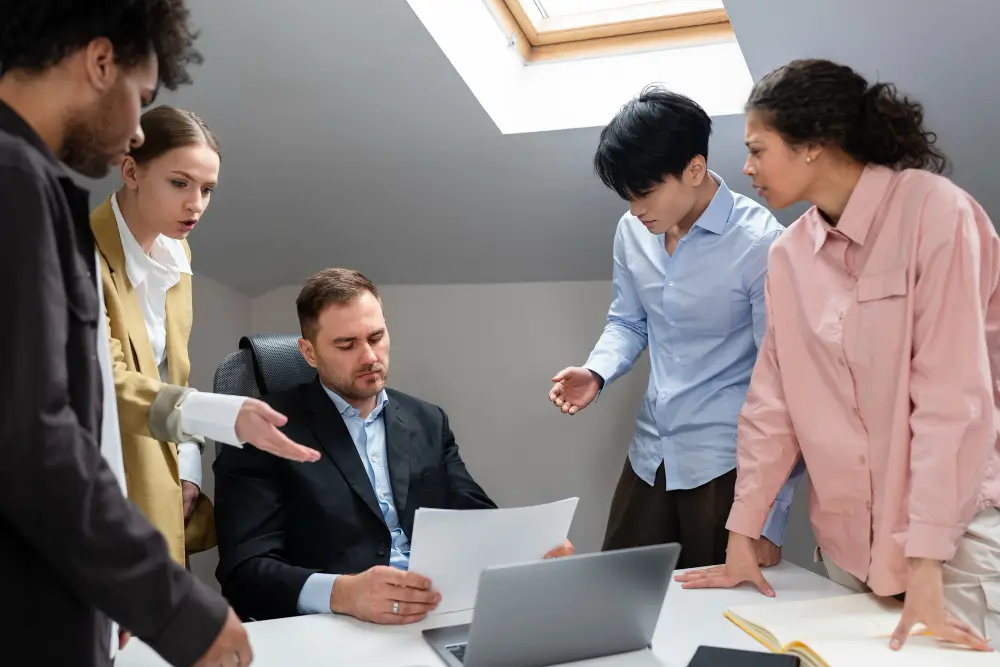 Team of five colleagues in an office discussing a document around a table, with a laptop open and papers in hand.