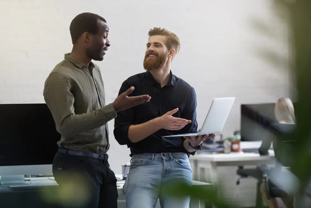 Two coworkers stand and chat in a bright office, one holding a laptop while gesturing to the other.